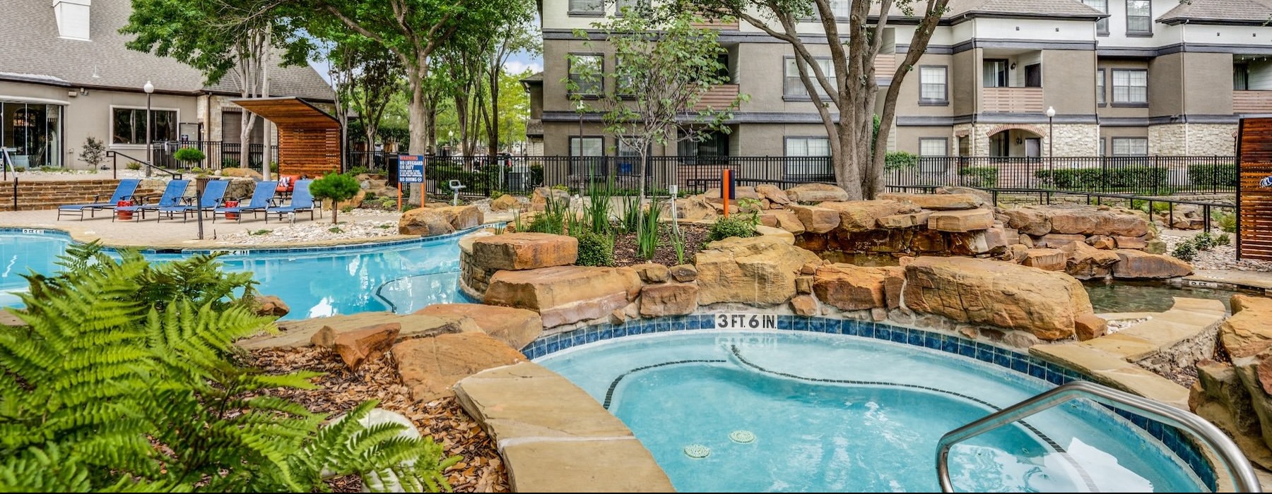 hot tub and pool with trees and rocks with a building behind it