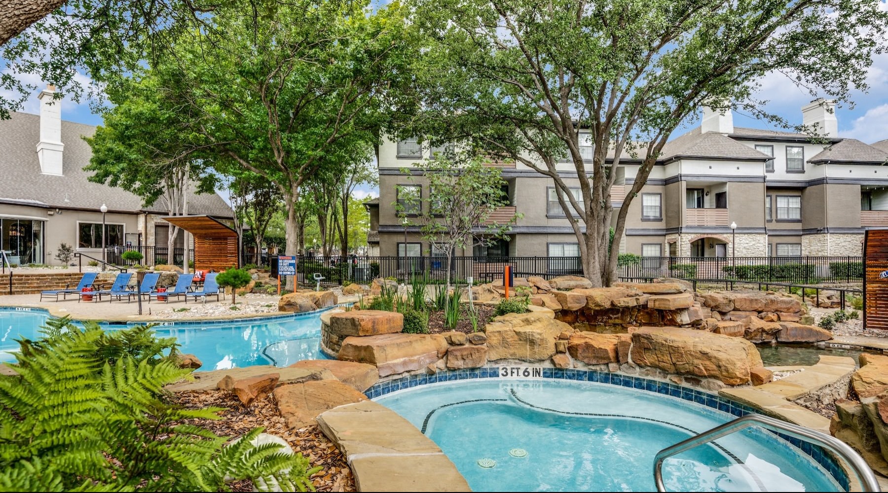 Resort-style swimming pool with lounge seating hot tub and pool with trees and rocks with a building behind it
